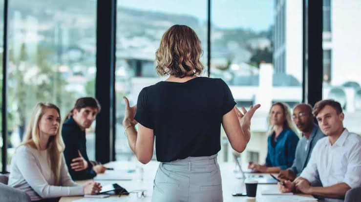Back View of a Woman Presenting to a Diverse Team Seated Around a Conference Table in a Bright Office.