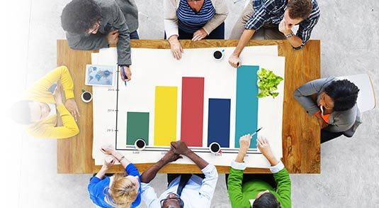 Team Assessment Team Building Zzeem Zzeem Overhead View of a Diverse Team Around a Table Analyzing a Colorful Bar Chart on a White Sheet.